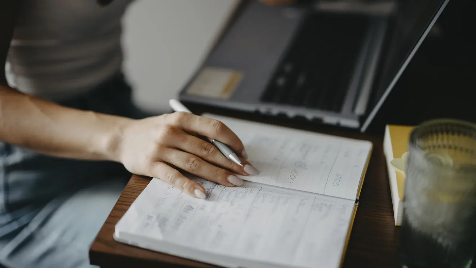 Person writing notes in a notebook beside an open laptop