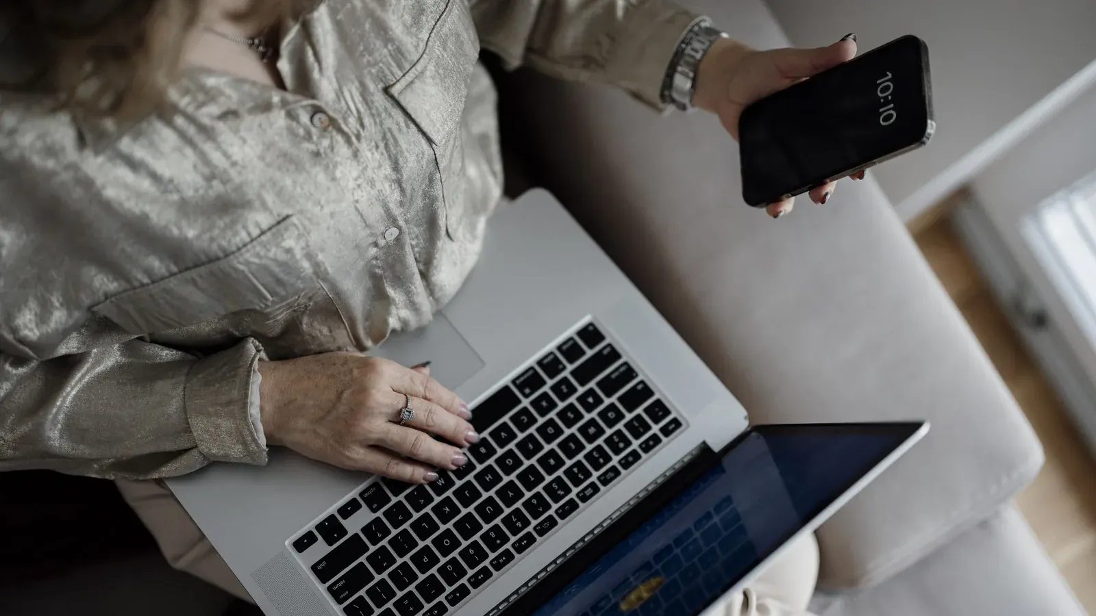 Woman sitting on a couch using a laptop while holding a smartphone